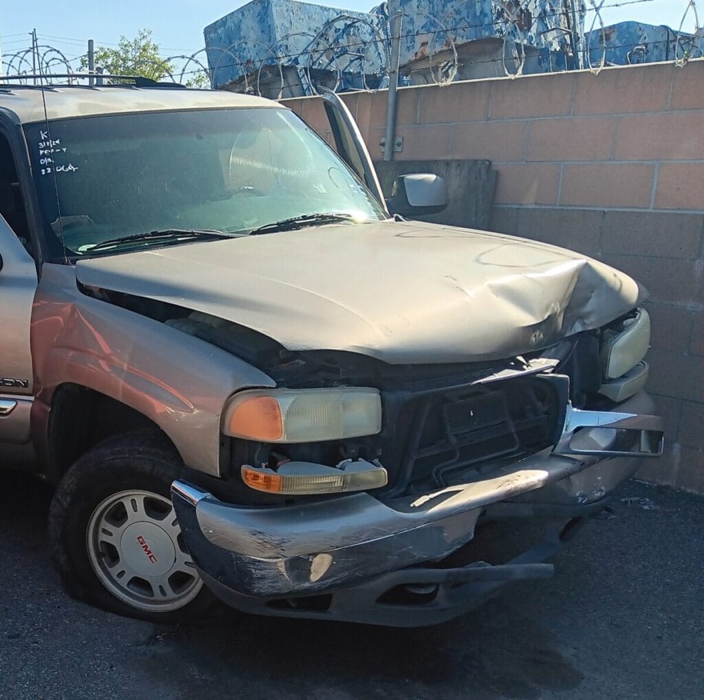 A severely damaged gold GMC Yukon SUV with a crumpled hood, a crushed front bumper, and a flat tire clearly with damaged radiator overheating