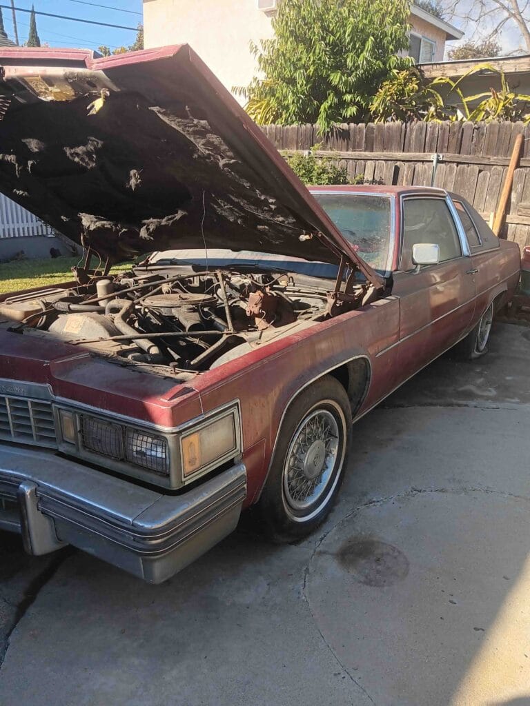 A dusty maroon 1970s classic Cadillac with the hood open and a flat tire parked in an Orange County driveway. Go Car Buyer pays cash for classic project cars in any condition.