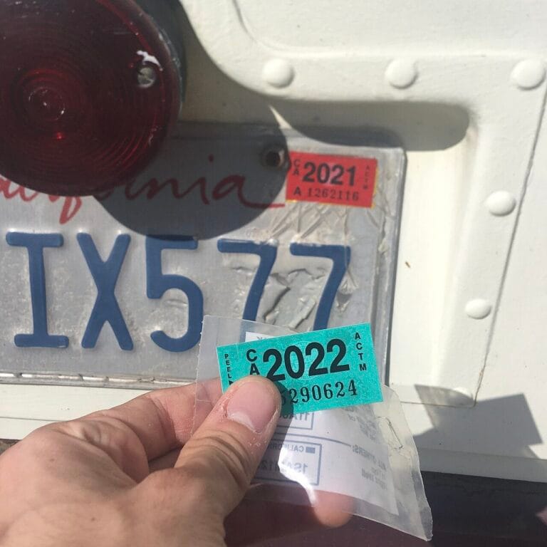 A hand holding a California DMV registration sticker next to a license plate with an expired tag.