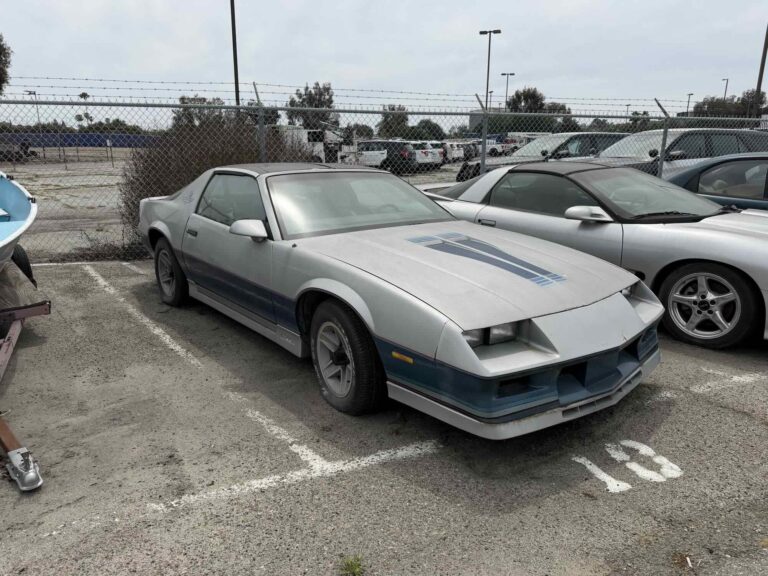 A dusty silver 1980s Chevrolet Camaro that has been sitting in a lot for years, representing a non-running vehicle.