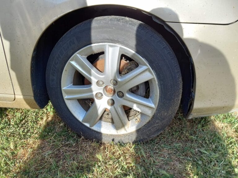 Close up of a car tire and silver alloy wheel parked on grass, showing a heavily rusted brake rotor behind the rim.